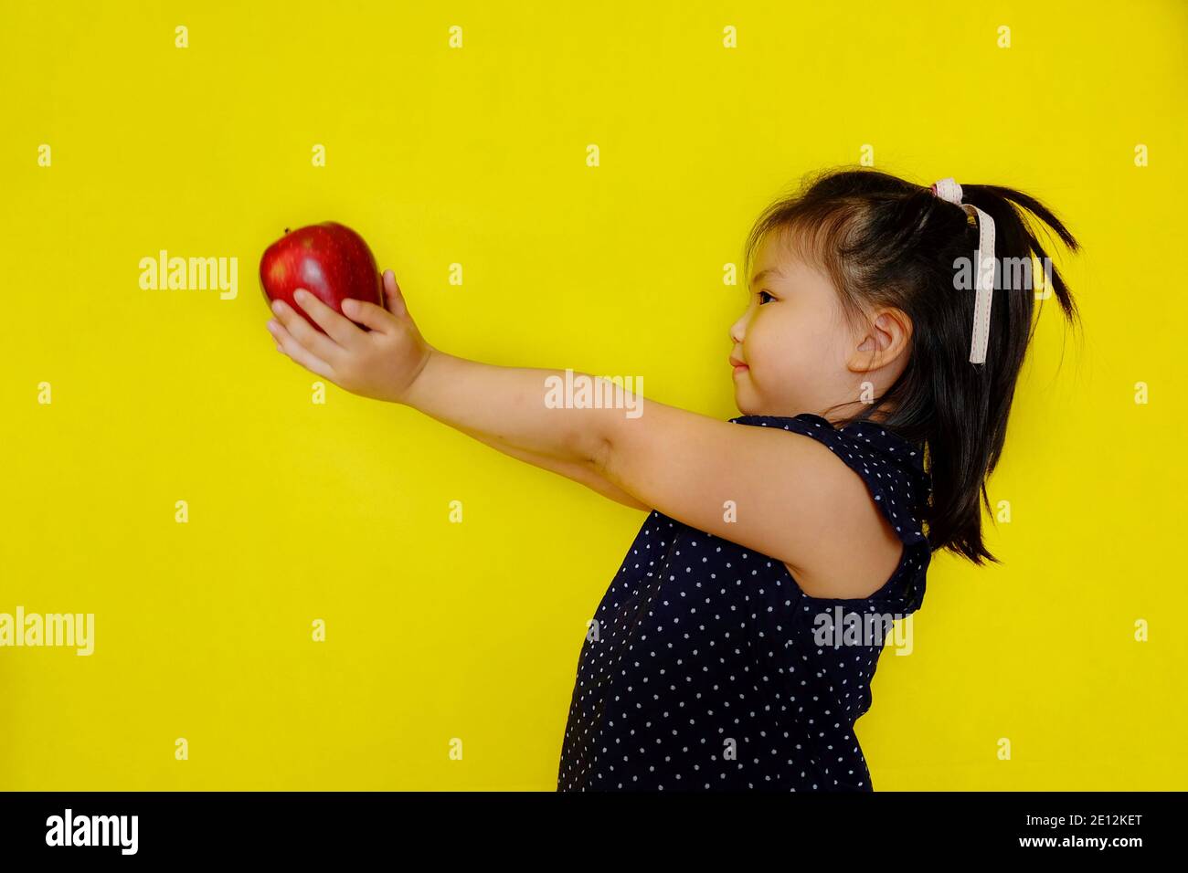 Una giovane ragazza asiatica carina a scuola, porgendo una mela rossa alla sua insegnante come regalo, sorridendo. Sfondo giallo brillante. Foto Stock