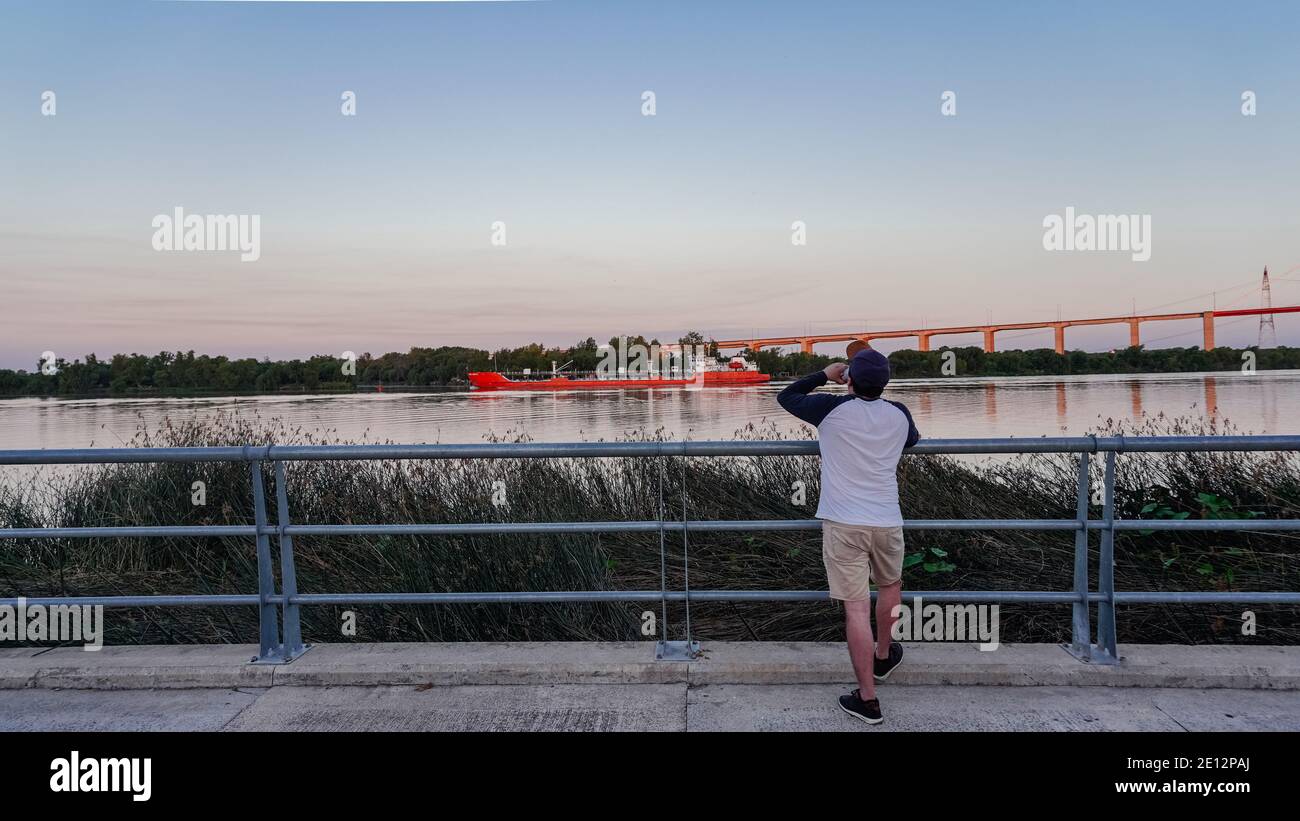 Uomo in piedi sulla vista posteriore di un uomo che guarda il ponte Zarate Brazo Largo ponte e il fiume parana, mentre una nave da carico attraversa di fronte, bere birra Foto Stock