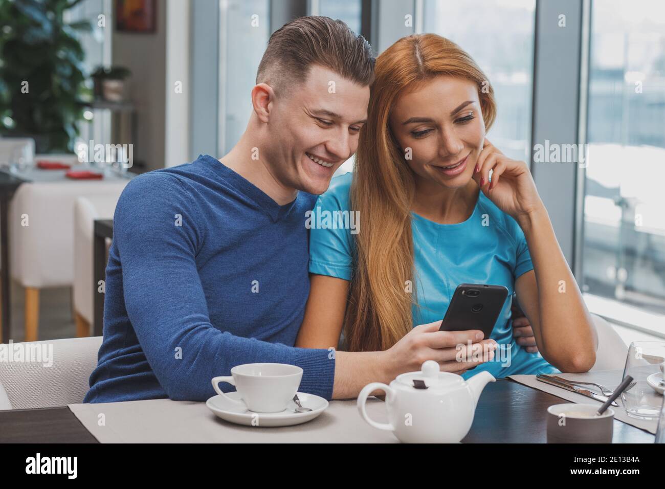 Bell'uomo sorridente, abbracciando la sua splendida ragazza, mostrandogli qualcosa sullo smartphone. Bella coppia che ha fatto colazione al caffè surf i Foto Stock