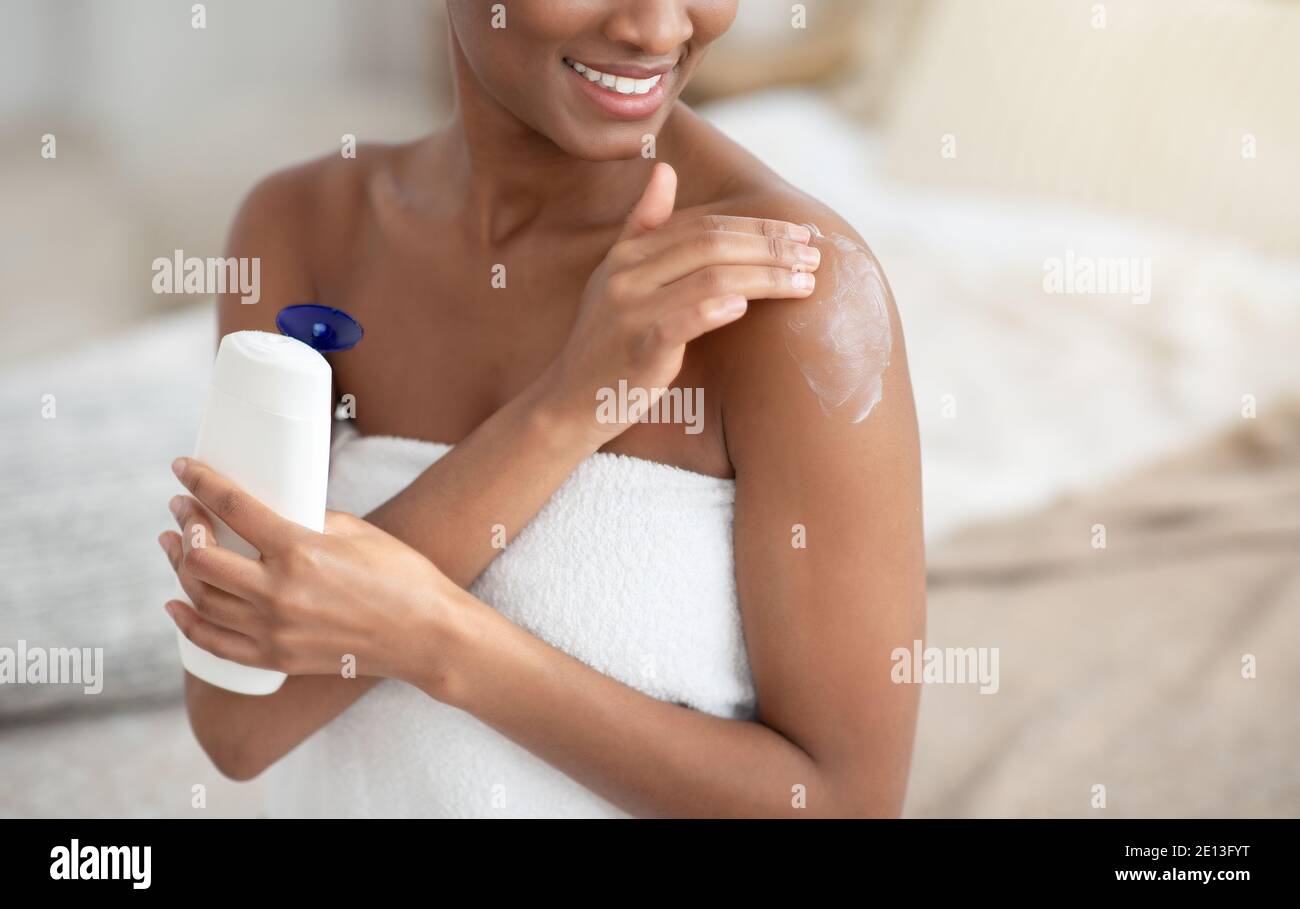 Felice donna afroamericana in asciugamano bianco dopo la doccia applica la crema al corpo Foto Stock