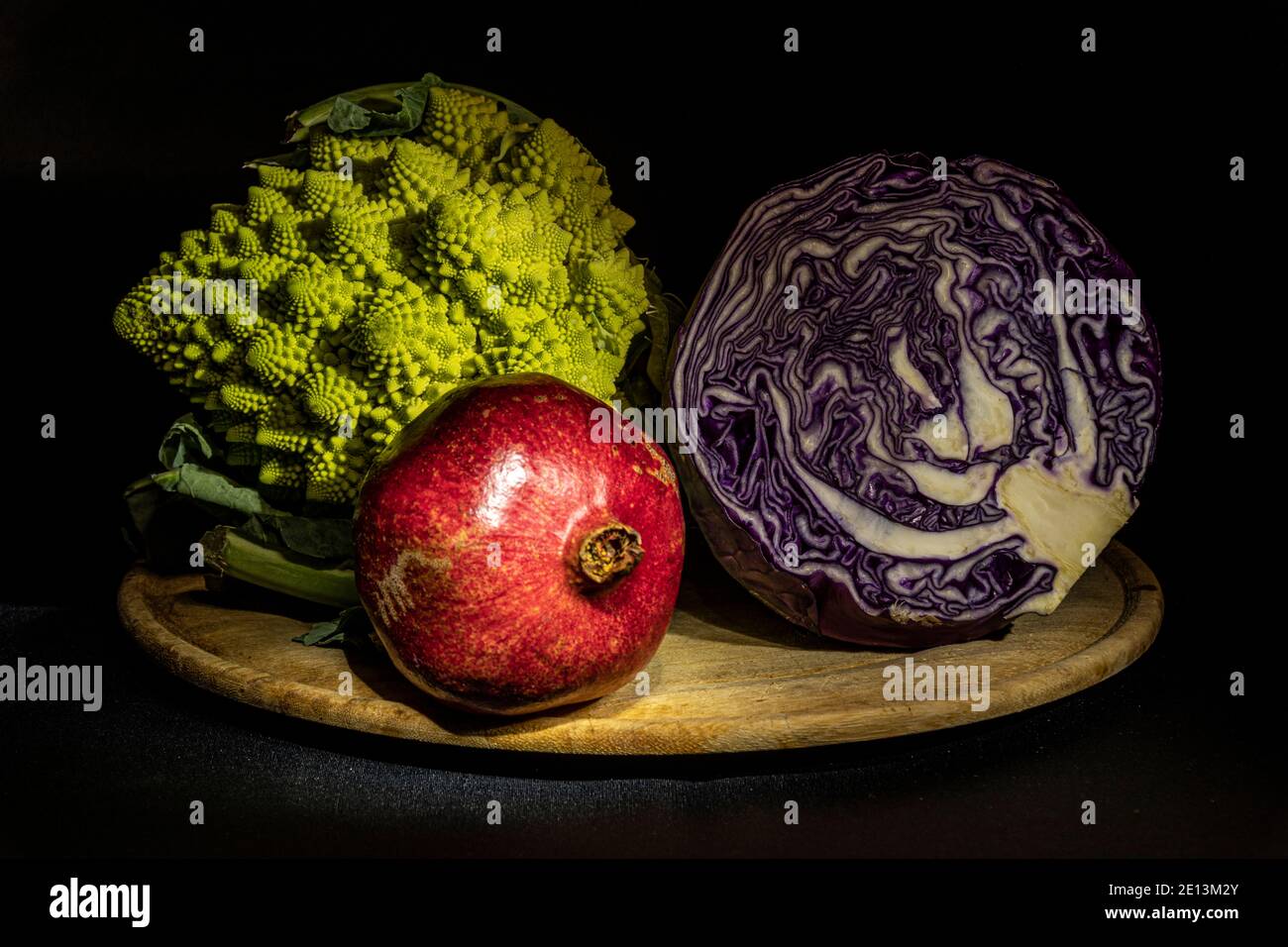 Verdure invernali. Composizione di natura morta con melograno, cavolo romano e crauti viola, su tagliere in legno su sfondo scuro. Foto Stock