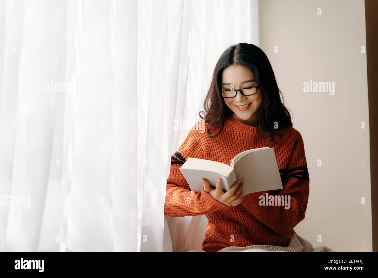 Donna asiatica felice in occhiali e maglione lettura libro e. guardando via con il sorriso mentre si riposa vicino alla finestra di casa Foto Stock