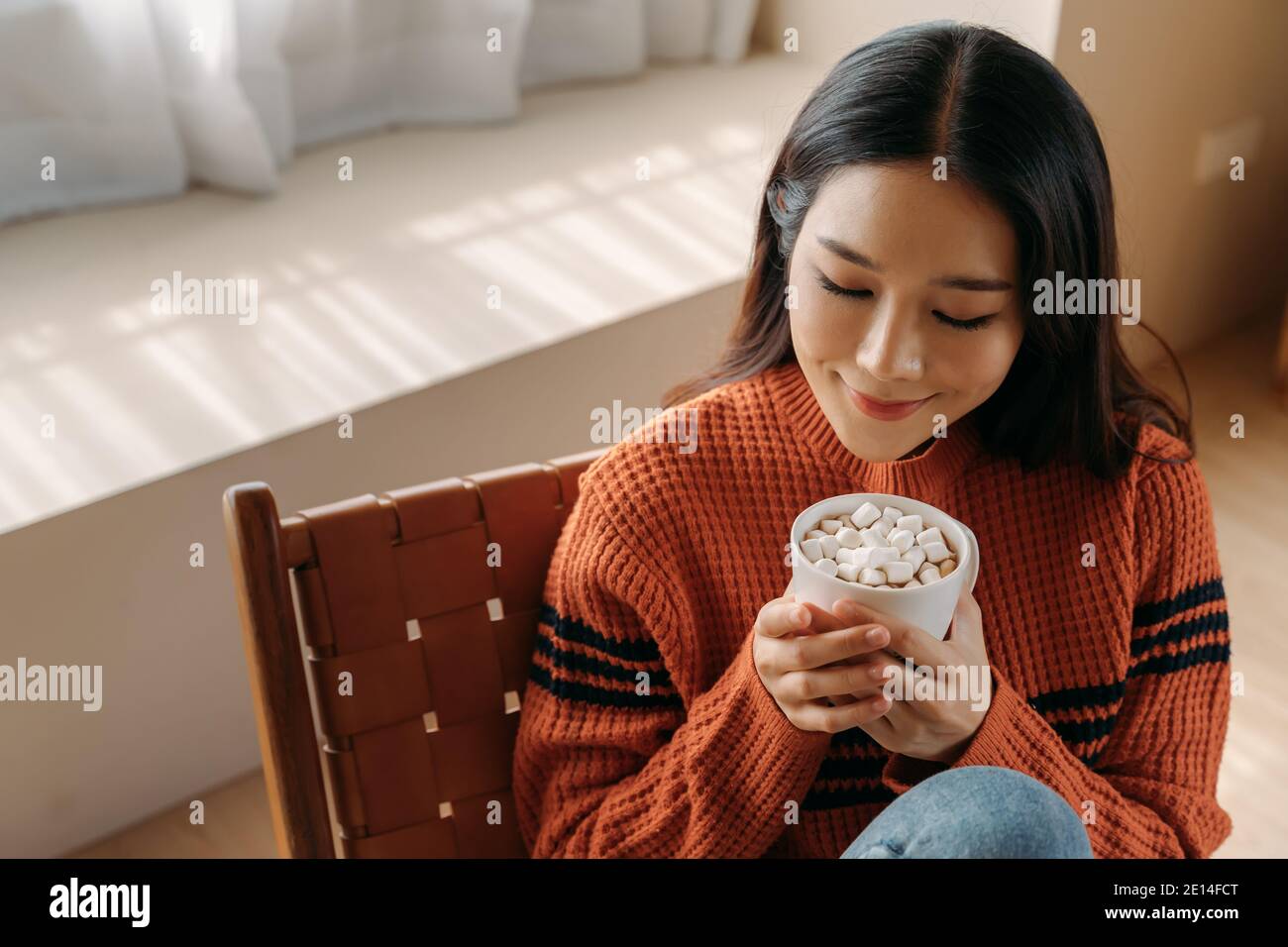 Felice giovane etnia femmina in maglione sorridente e godendosi caldo bevanda con marshmellow mentre riposa su una comoda sedia a casa Foto Stock