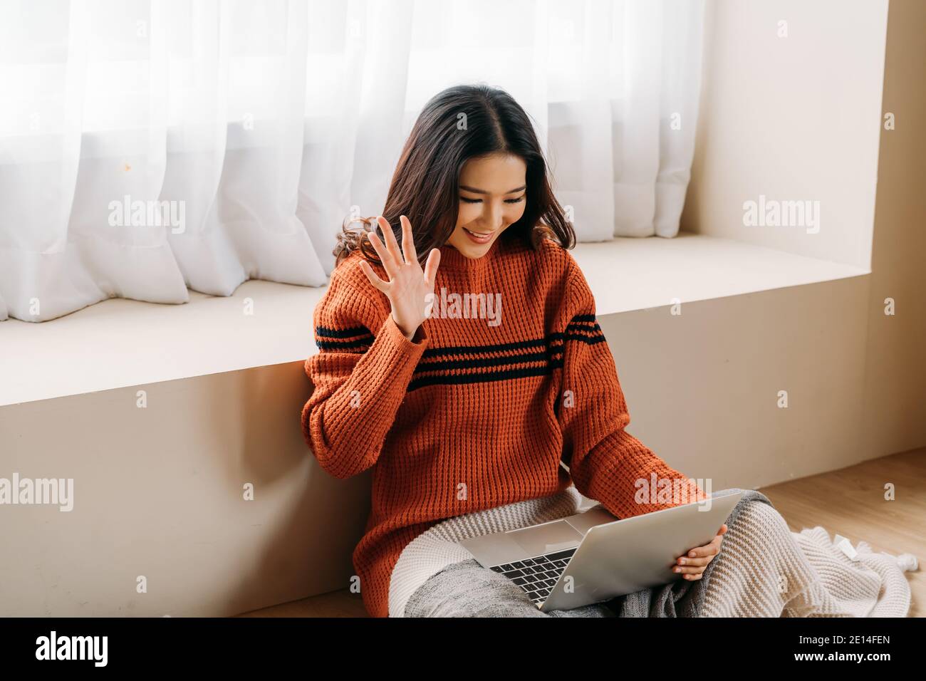 Felice donna etnica in caldo maglione sorridente e ondeggiante mano mentre si è seduti a terra e si effettua una videochiamata sul computer portatile Foto Stock