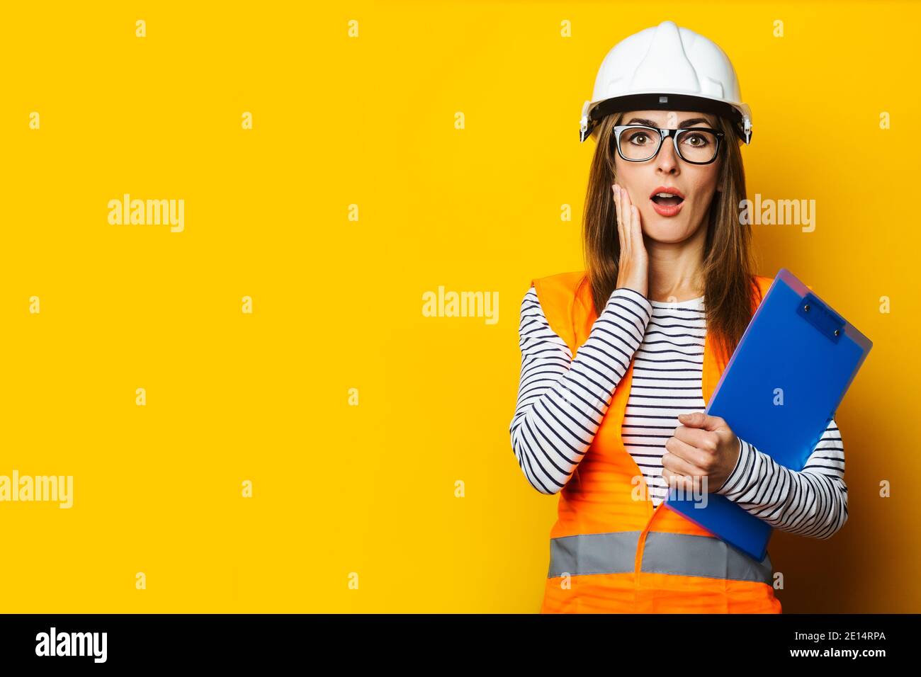 Giovane donna con un volto sorpreso in un gilet e elmetto tiene una clipboard su uno sfondo giallo. Concetto di costruzione, nuovo edificio. Banner. Foto Stock
