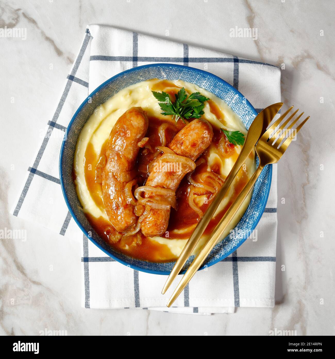 Bangers di cibo britannico e mash o salsiccia di maiale con sugo di cipolla e purè di patate su un piatto con posate dorate su un tavolo di marmo, vista dall'alto, primo piano Foto Stock