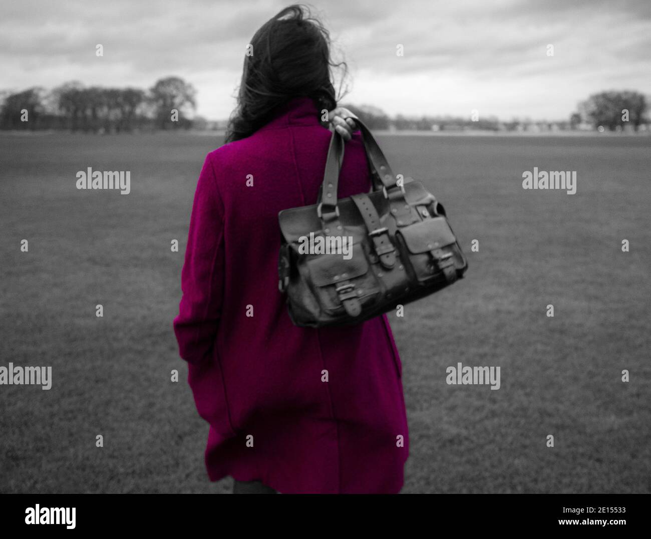 Vista posteriore con messa a fuoco morbida della donna che tiene la borsa in pelle Mulberry Roxanne sopra le spalle nel parco. Colore desaturato con pop di colore magenta Foto Stock