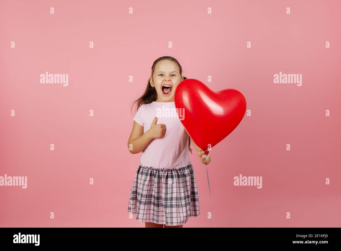 primo piano entusiasta rapturous estatica ragazza in abito rosa tiene rosso palloncino a forma di cuore e dà pollici in su isolato su sfondo rosa Foto Stock