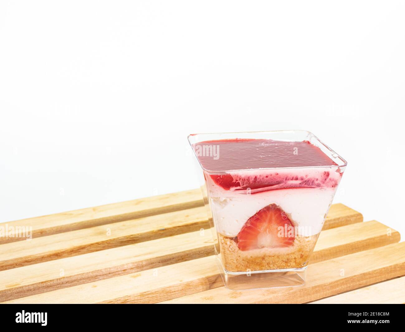 Primo piano della cheesecake alla fragola in una tazza trasparente di plastica quadrata su tavola di legno su sfondo bianco. Foto Stock