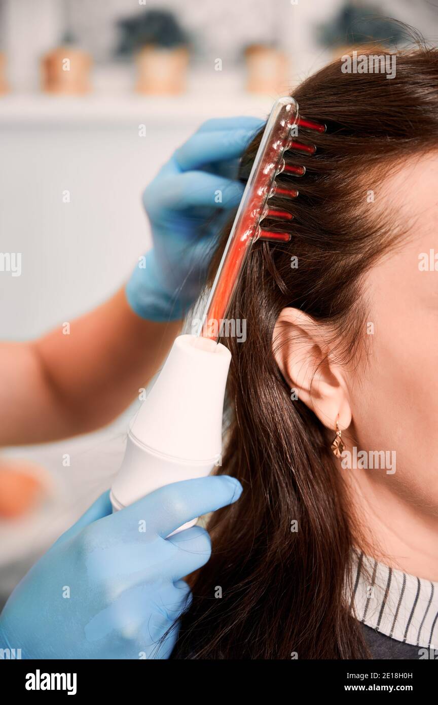 Primo piano della mano del medico tricologista in guanti sterili  spazzolando i capelli del cliente femminile con il dispositivo laser di  crescita dei capelli. Donna che riceve trattamento dei capelli terapia laser .
