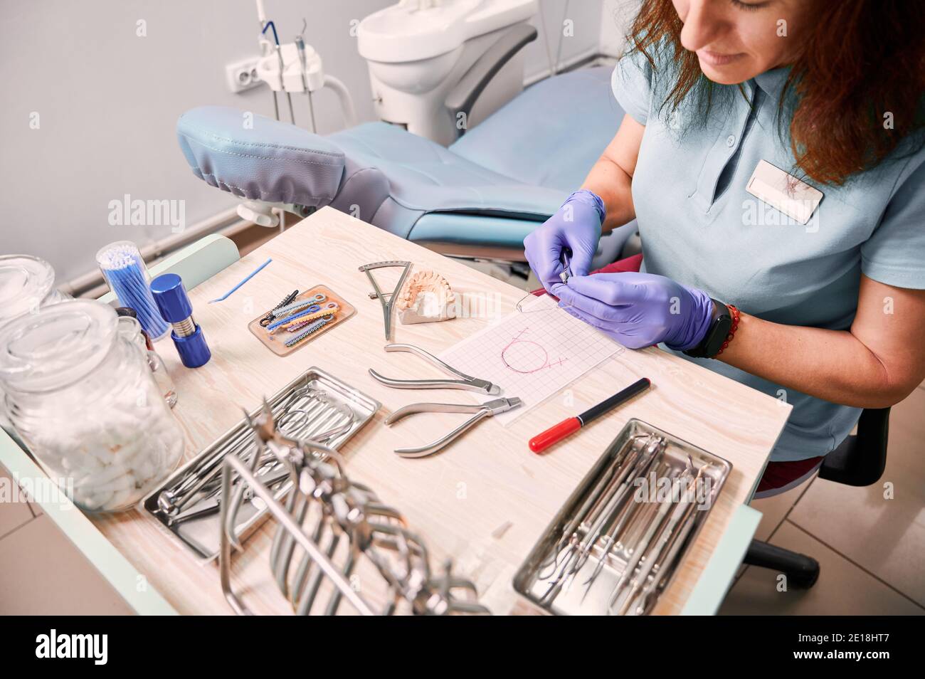 Vista dall'alto del dentista femminile in guanti sterili, mentre si siede al tavolo con strumenti e strumenti ortodontici. Ortodontista donna che prepara il filo per l'attacco dei rinforzi. Foto Stock