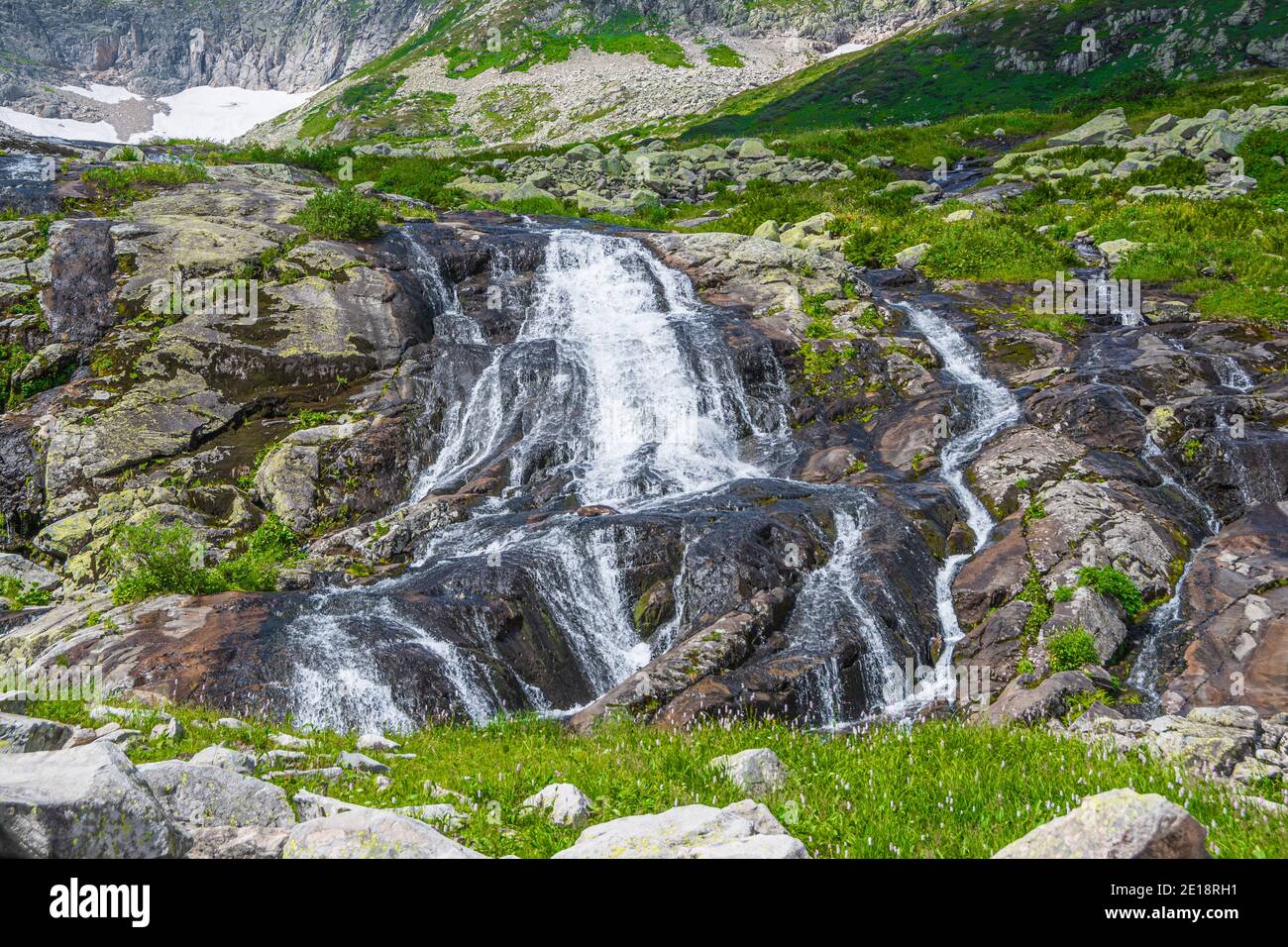 Rapido ruscello in valle di montagna tra le rive erbose. Piccola cascata in prato verde. Foto Stock