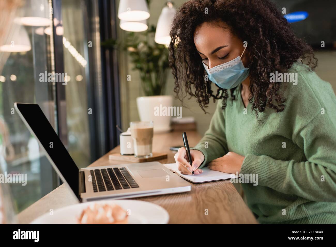 donna afroamericana in maschera medica di scrittura in notebook vicino computer portatile con schermo vuoto sul tavolo Foto Stock