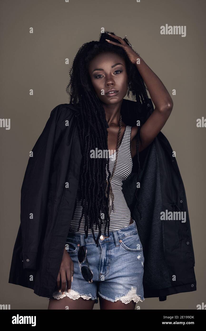 Ritratto di una giovane femmina nera con lunghi dreadlock, bel trucco e labbra morbide che si posano da sola indossando gioielli e pantaloncini in denim. Foto Stock