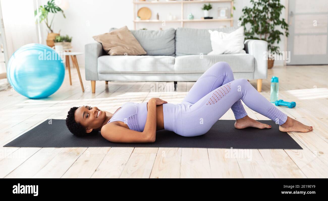 Giovane afroamericana sdraiata sul tappeto yoga in soggiorno, riposandosi dopo un intenso allenamento, panorama Foto Stock