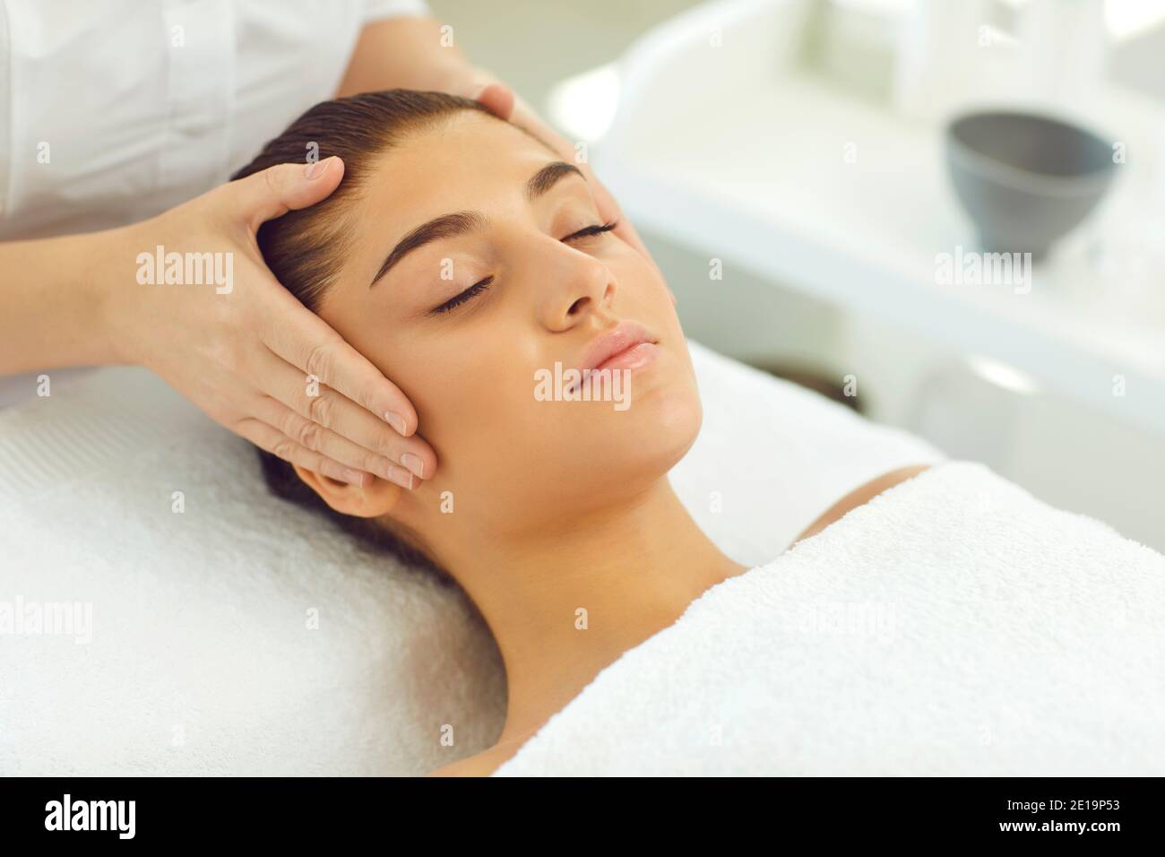 Mani di cosmetologist che toccano la testa e che controllano la pelle o che fanno massaggio viso per giovane donna Foto Stock