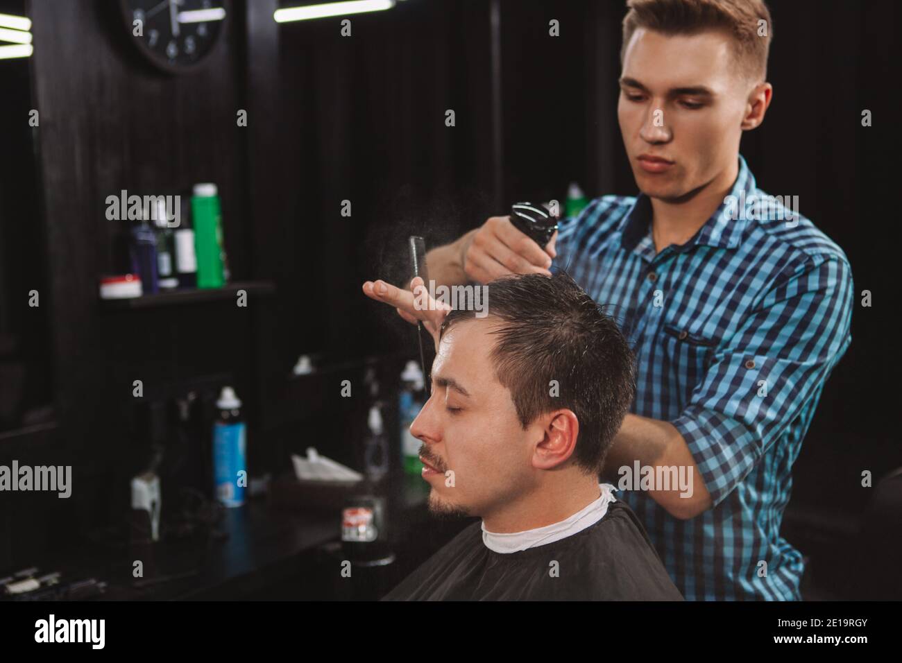 Giovane barbiere attraente che lavora con un cliente, spruzzando i capelli con acqua prima di dare un taglio di capelli. Uomo maturo al barbiere locale Foto Stock