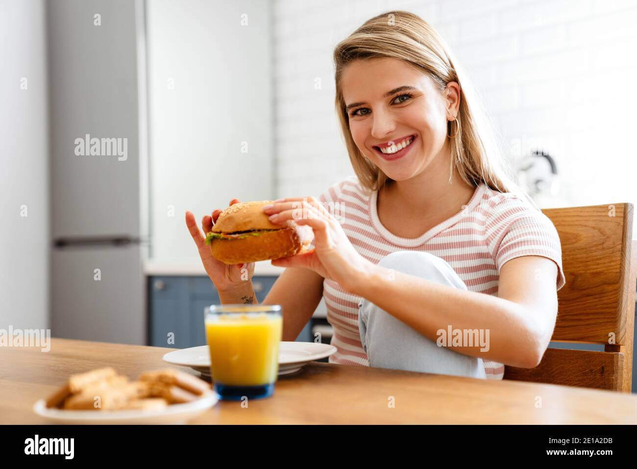 Sorridente giovane donna che mangia hamburger in cucina a casa Foto Stock