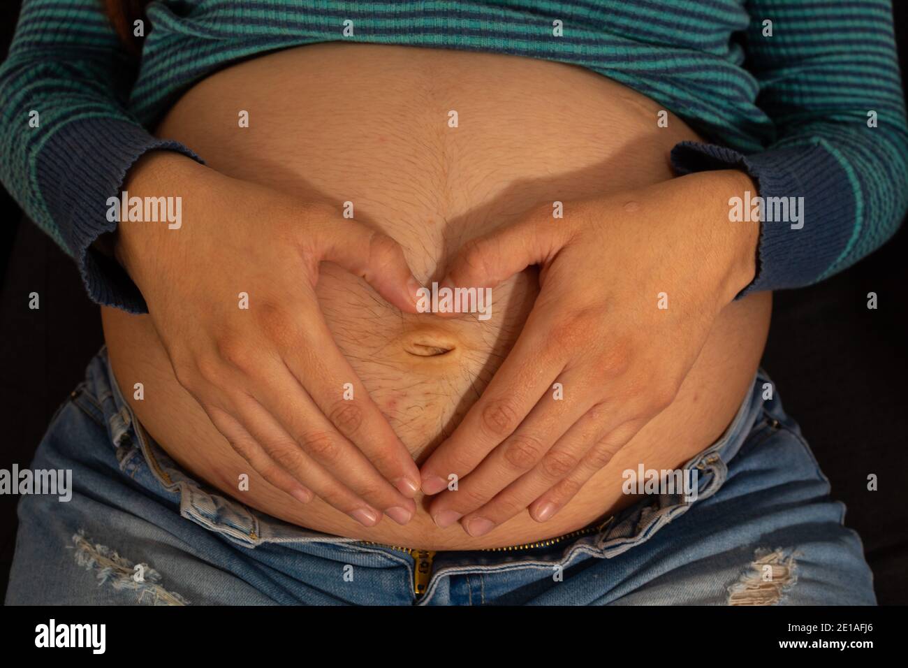 primo piano del ventre di una donna incinta dentro il suo terzo trimestre indossando abiti casual che fanno un cuore con le mani Foto Stock