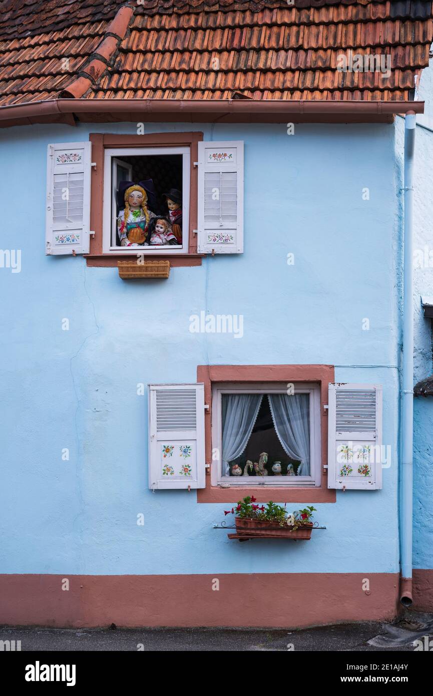 Varie bambole dipinte e vestite puntano attraverso finestre decorate di una casa blu chiaro. Foto Stock