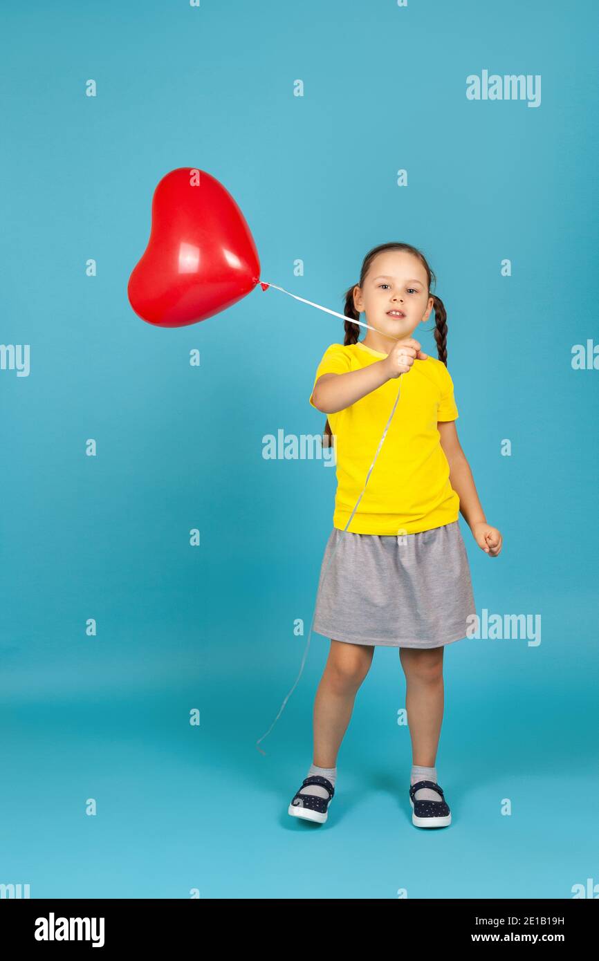 Ragazza a tutta lunghezza con i pigtail in T-shirt gialla e gonna grigia danze e onde un palloncino rosso a forma di cuore, isolato su uno sfondo blu Foto Stock