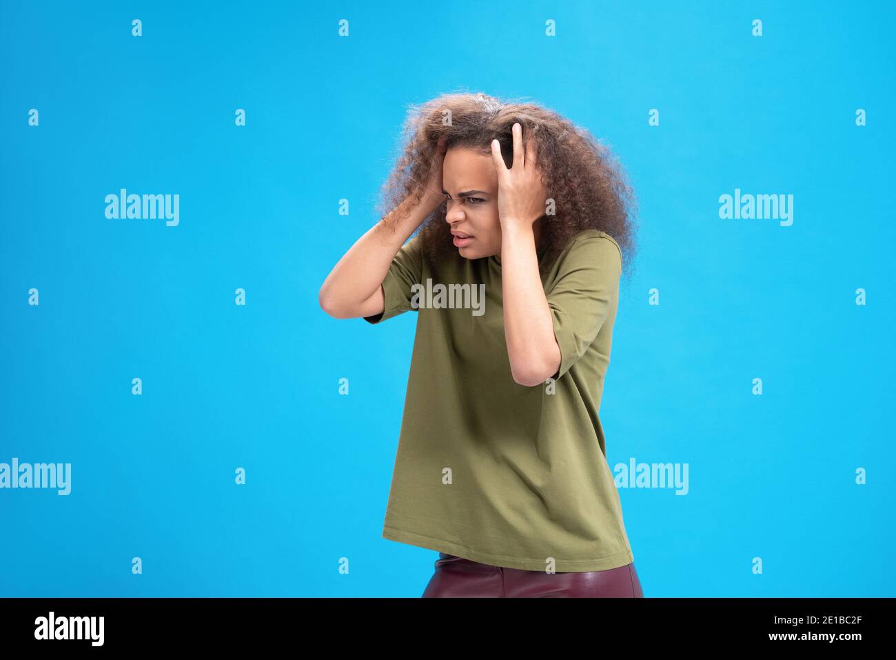 Esperienza di emicrania o mal di testa giovane donna afroamericana in piedi tenendo testa con le mani in t-shirt oliva isolato su sfondo blu. Umano Foto Stock