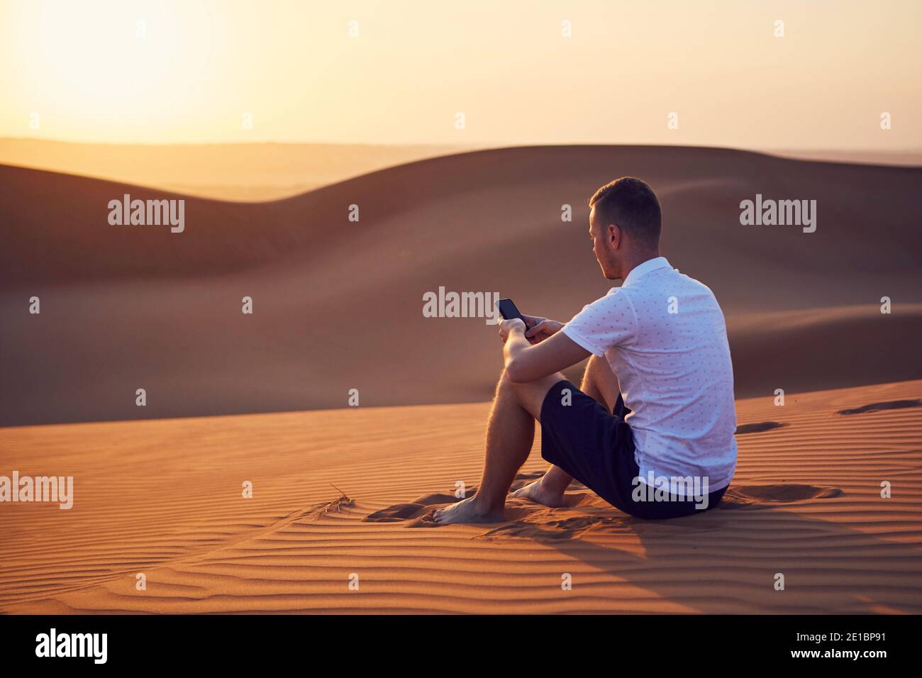Solo nel deserto. Giovane uomo seduto sulla duna di sabbia e utilizzando il telefono al tramonto dorato. Wahiba Sands in Oman. Foto Stock