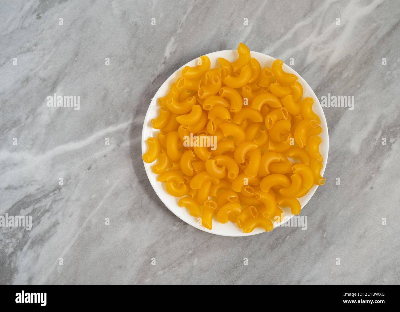 Maccheroni a gomito organici su una piastra bianca in cima a un piano di appoggio grigio illuminato con luce naturale. Foto Stock