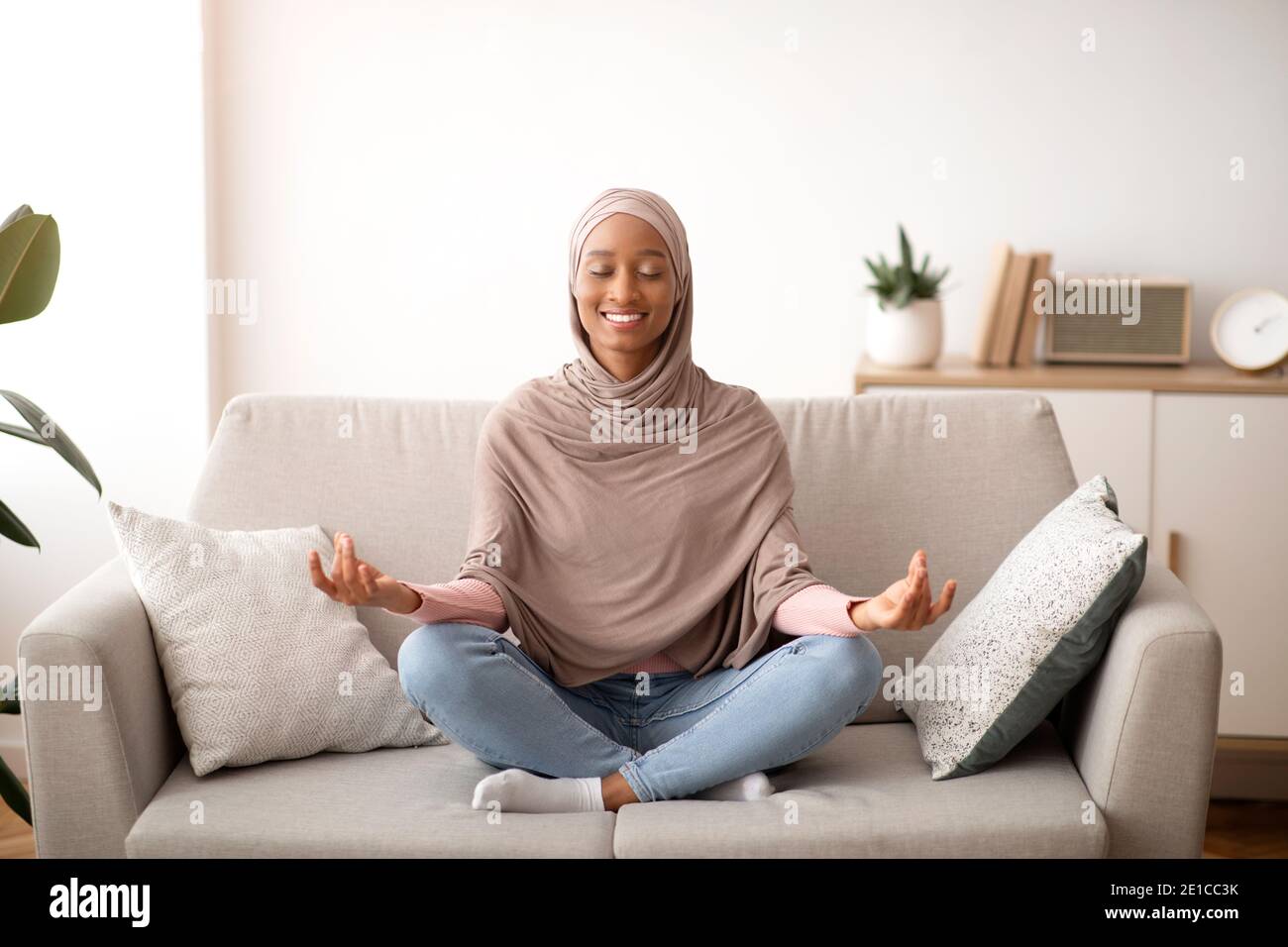 Concetto di gestione dello stress. Giovane donna tranquilla in hijab meditando con gli occhi chiusi sul divano a casa Foto Stock