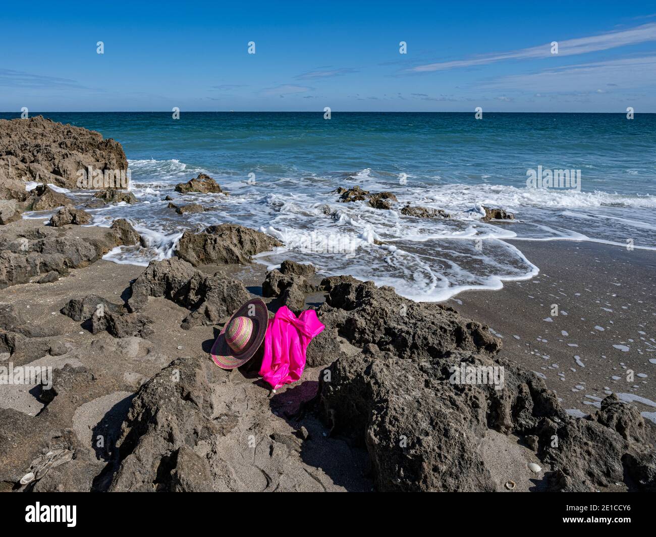 Sfondo di stagcape con un vestito da bagno rosa caldo che giace sulle rocce con un cappello di paglia mentre le onde dolci spruzzano e fluiscono sopra la sabbia e le rocce di un . Foto Stock