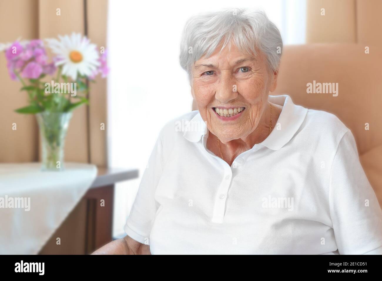 Donna anziana sorridente con una stoop osteoporosi in una sedia comoda nella sua stanza ad una casa di riposo. Foto Stock