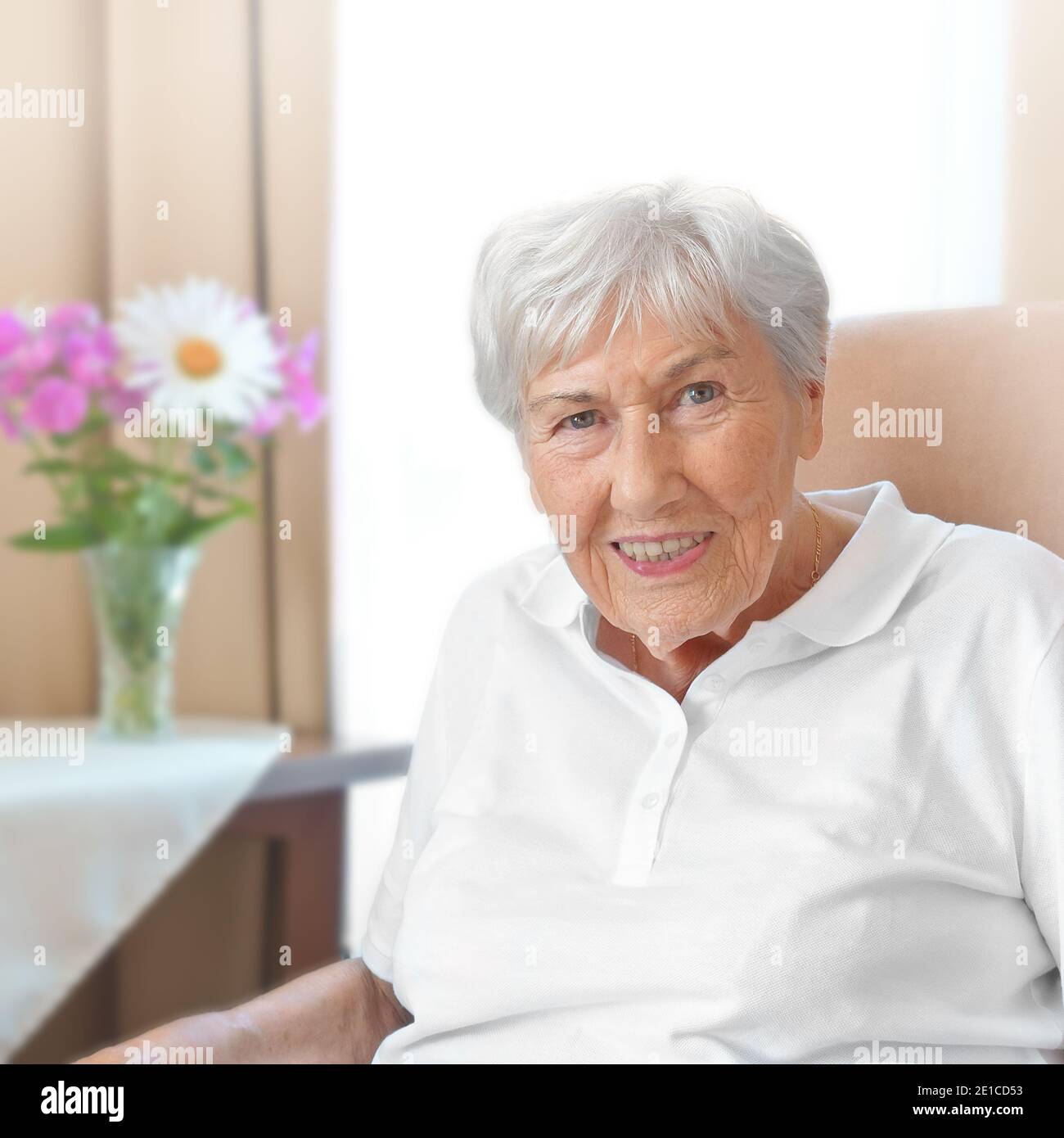 Donna anziana sorridente con una stoop osteoporosi in una sedia comoda nella sua stanza ad una casa di riposo. Foto Stock