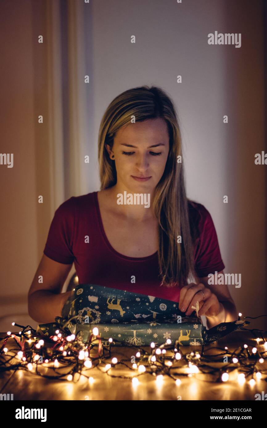 Ragazza di etnia europea con i capelli biondi con interesse savvolge un regalo per Natale o compleanno nella calda luce di piccole luci. Vacanze, benessere Foto Stock