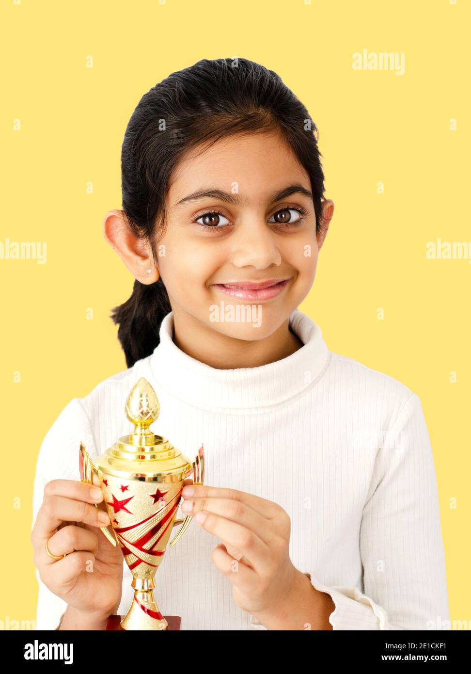 Ritratto di una bella e sorridente scuola indiana ragazza su sfondo giallo con un trofeo d'oro e raffigurante il concetto vincente. Foto Stock