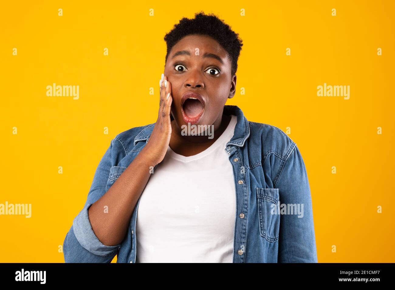 Shocked Plus-Size African Woman guardando fotocamera in posa, sfondo giallo Foto Stock