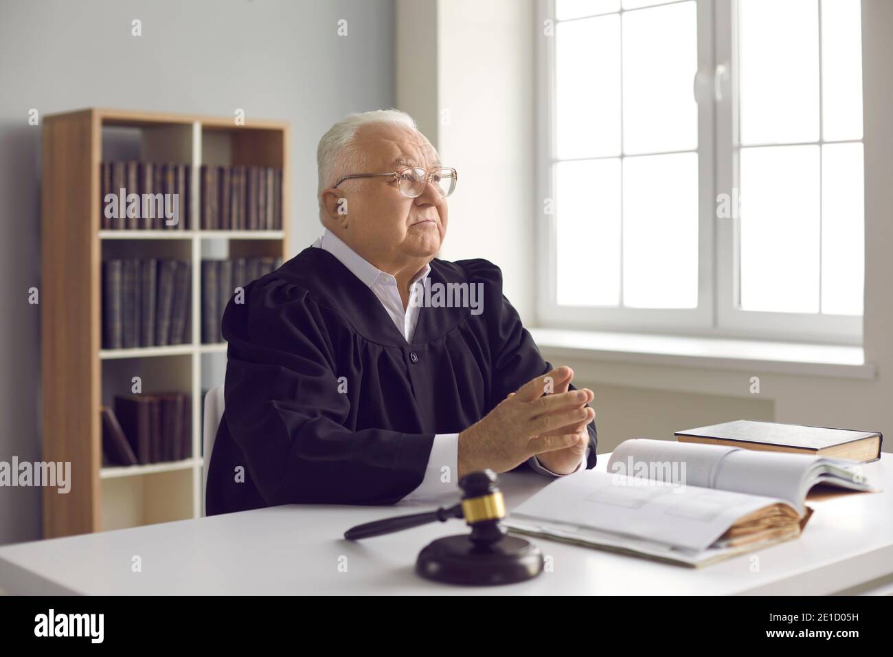 Giudice serio maturo seduto alla scrivania con libro di legge aperto nel suo ufficio o in aula Foto Stock