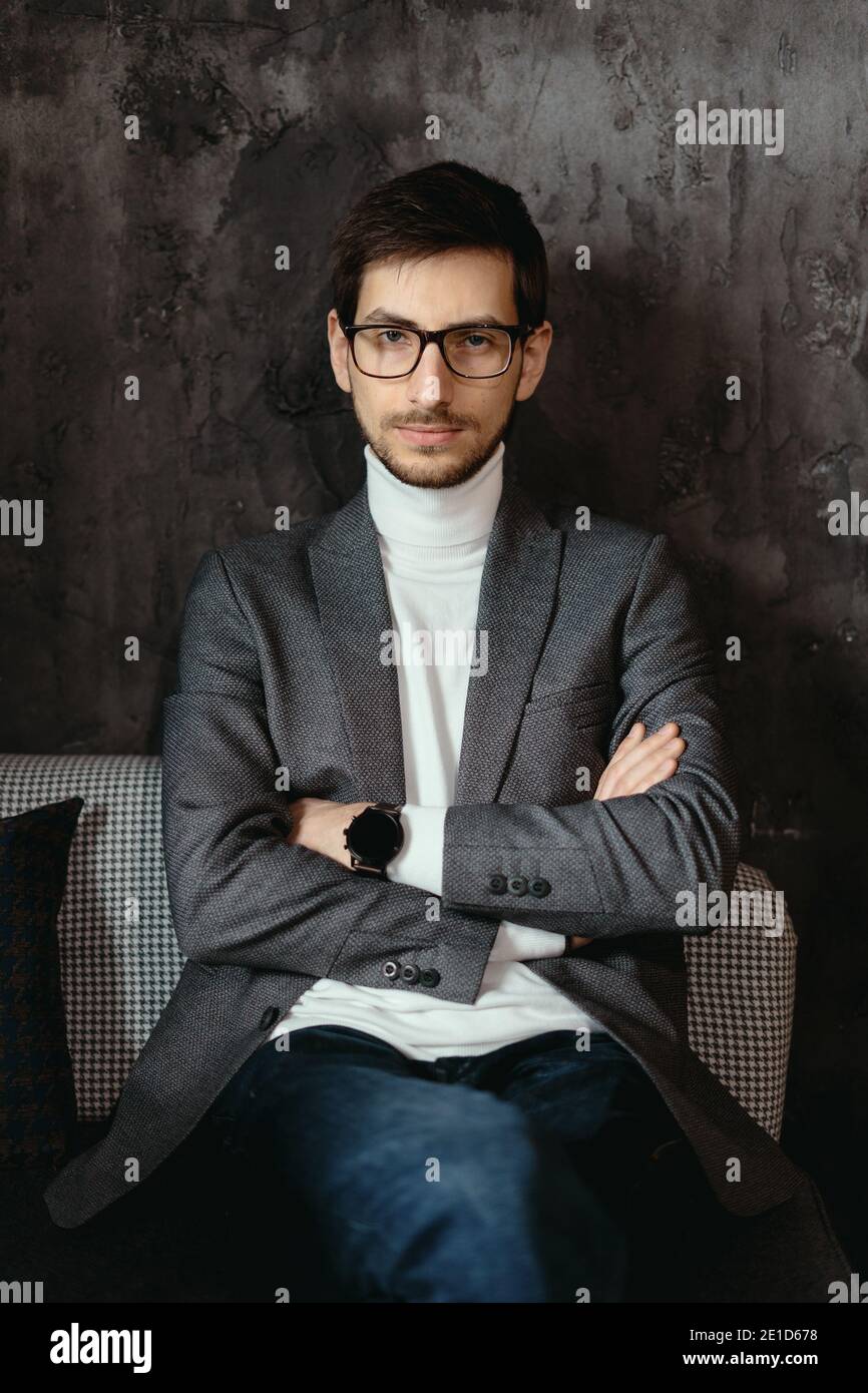 Giovane uomo d'affari di successo che indossa occhiali, giacca grigia, dolcevita bianco su sfondo grigio scuro Foto Stock