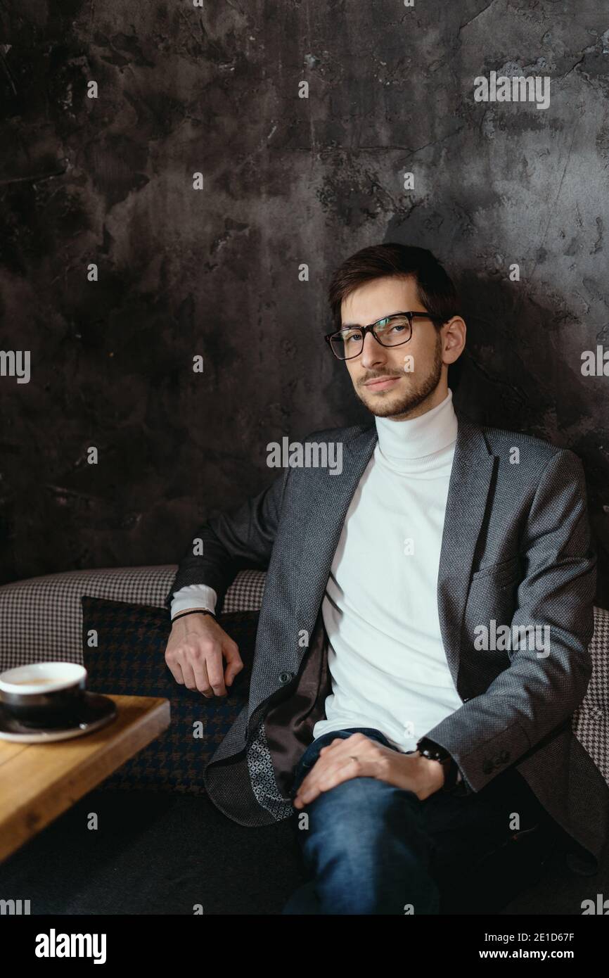 Giovane uomo d'affari di successo che indossa occhiali, giacca grigia, dolcevita bianco su sfondo grigio scuro Foto Stock