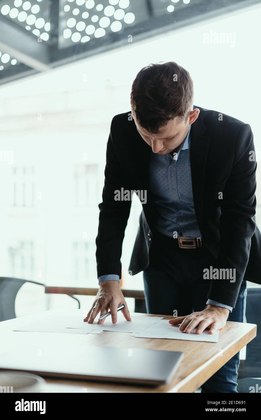 Giovane uomo d'affari sicuro che firma i documenti in un ufficio moderno con la finestra sullo sfondo. Penna in mano, carte sulla scrivania di legno Foto Stock