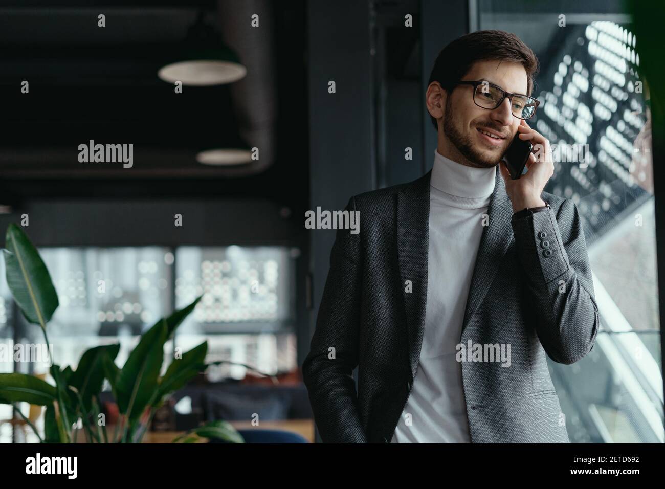Un carismatico giovane uomo d'affari che indossa occhiali, giacca grigia, dolcevita bianco che ha una conversazione al telefono in uno spazio moderno Foto Stock