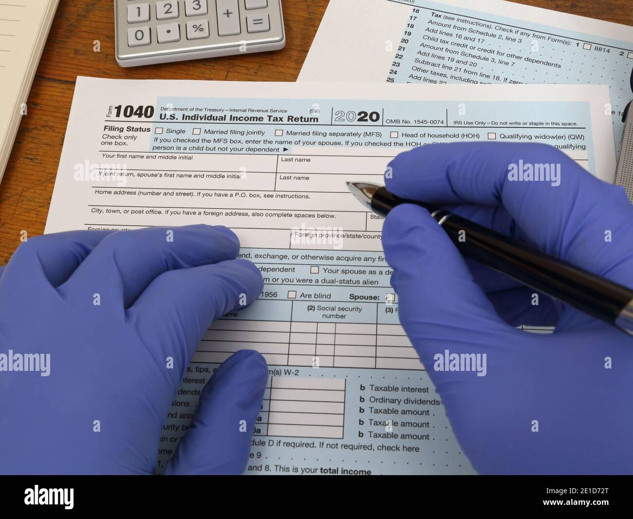 Le mani che indossano i guanti di gomma di nitrile blu sono mostrate iniziando a compilare un modulo fiscale statunitense 1040, rilasciato dal Dipartimento del Tesoro e IRS. Foto Stock