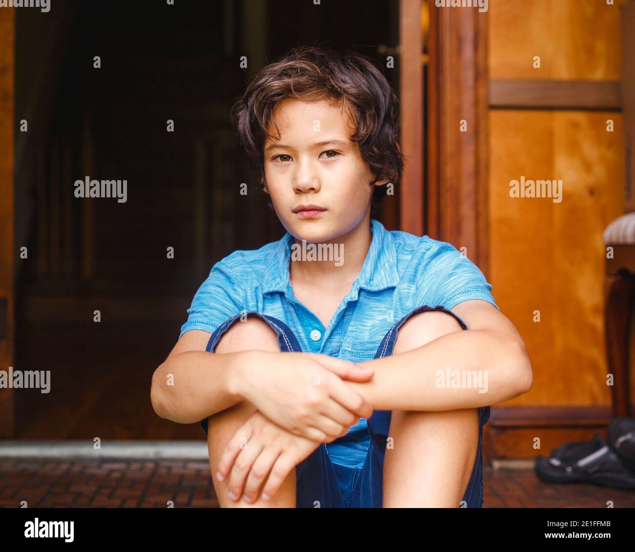 Un ragazzo con pelle dorata ed espressione seria si siede dentro porta di casa Foto Stock