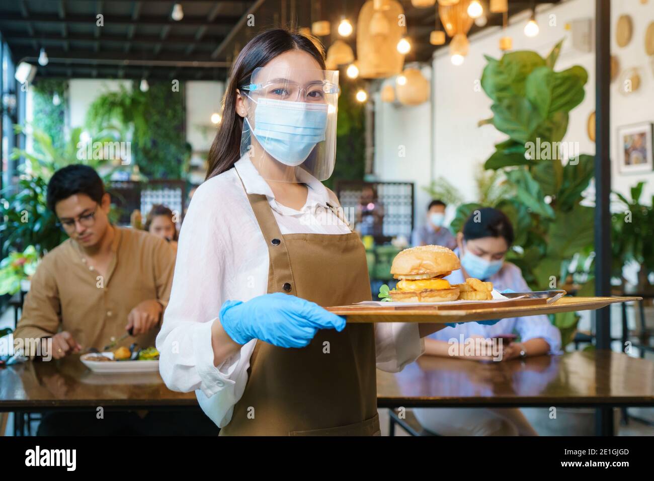 Ritratto attraente cameriera asiatica indossare maschera viso e visiera di protezione tenendo vassoio cibo per servire il pasto al cliente con il custode in background. Nuovo normale Foto Stock
