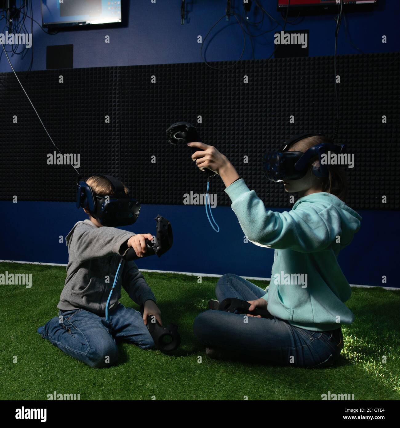 due bambini che indossano cuffie per realtà virtuale e con joystick giocano videogiochi seduti nell'area giochi Foto Stock