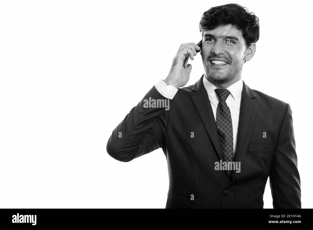 Riflessivo felice giovane imprenditore persiano sorridente mentre parlano al telefono cellulare e guardando in alto Foto Stock