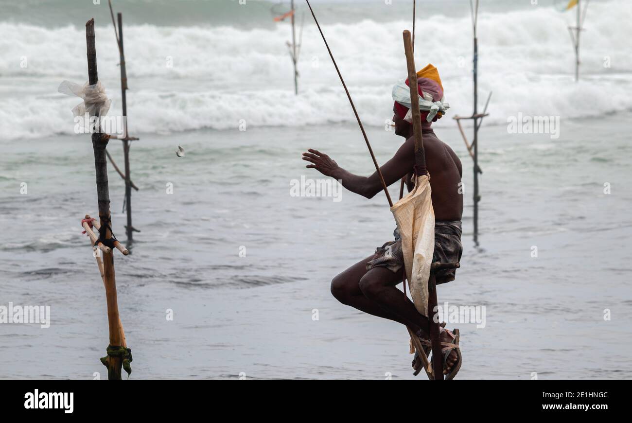 Weligama, Sri Lanka - 07 26 2020:Vecchio pescatore che tiene una rode in un palo di legno mentre si siede, pesca la sera, cattura un piccolo pesce, una ricompensa per Foto Stock