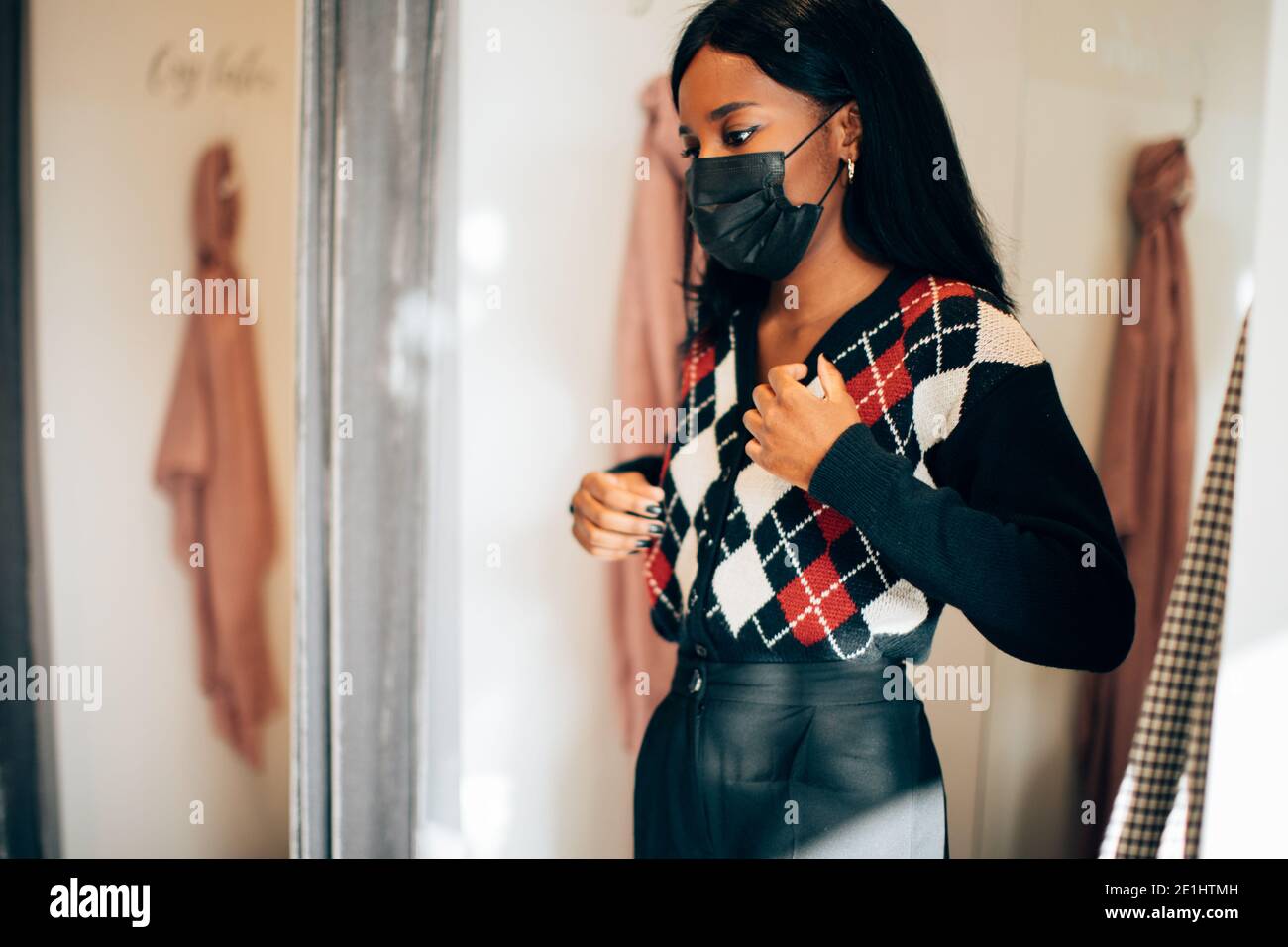 giovane donna con una maschera nera in piedi in un negozio di moda che indossa un abito mentre si guarda nel speculare Foto Stock