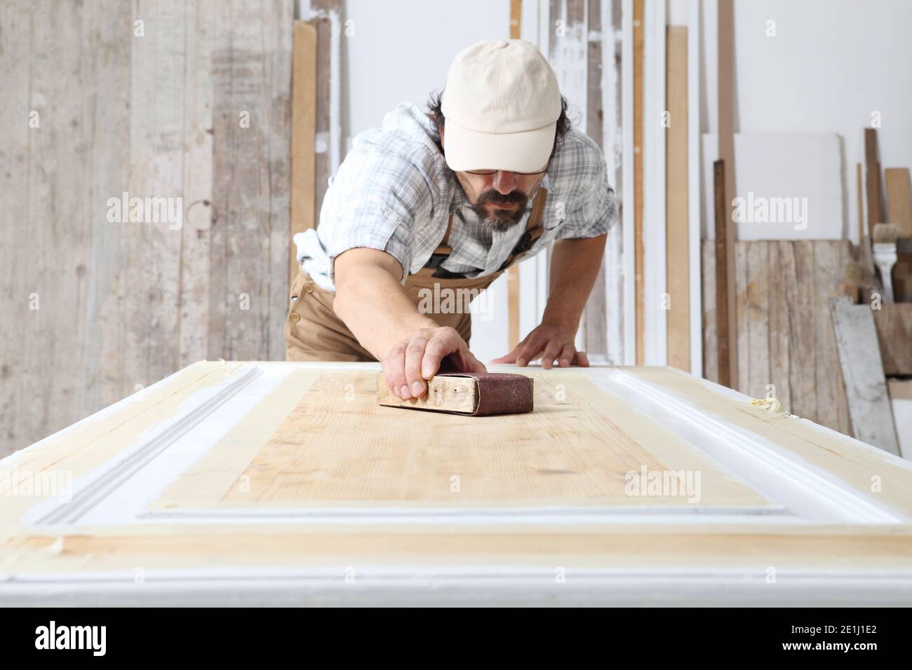 Falegname maschio che lavora il legno in officina di Falegnameria, levigando una porta di legno con carta vetrata, indossando tuta e cappuccio Foto Stock