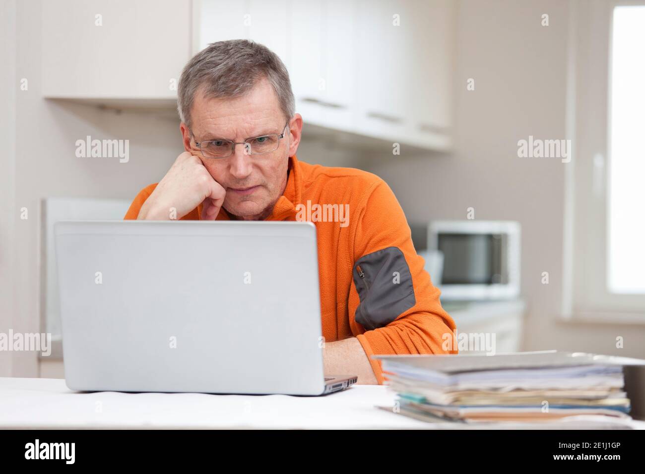 Uomo pensivo maturo che lavora a distanza da casa con il laptop e. files - sullo sfondo una cucina luminosa Foto Stock