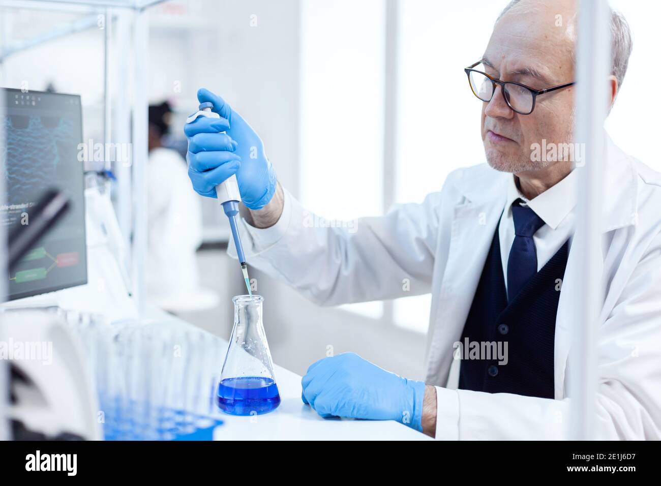 Scienziato esperto che lavora in laboratorio biotecnologico con contagocce sterile e liquido blu. Chimico professionista senior che utilizza pippete con soluzione blu per test microbiologici. Foto Stock